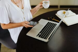 Woman sitting at a table drinking tea while the other hand on her phone with a laptop open on her table